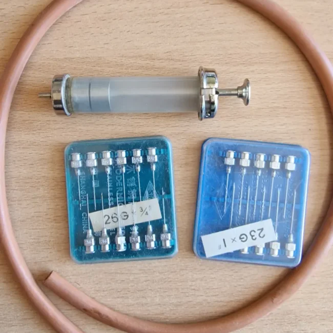 Instruments — contents of the bag. A reusable glass-and-chrome syringe, a length of rubber tubing, and two trays of stainless-steel needles (26G ¾" and 23G 1"). The minimal kit of a state-employed midwife mid-century